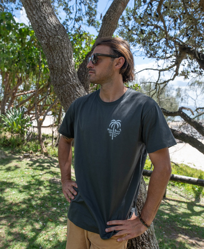 A man in the Golden Breed Manly Palm Loco Tee (Wash Black) and sunglasses stands outdoors by a tree, with lush greenery and Manly Beach in the background.