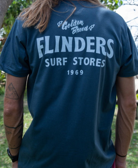 Person outdoors wearing the Golden Breed Flinders Easy Rider Tee | Denim, a 100% Cotton Jersey with Golden Breed FLINDERS SURF STORES 1969 on the back, part of our Location Tees collection.