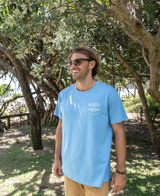 A man in Golden Breed’s Manly Boards Tee | Azure Blue stands outside on grass near trees, smiling and glancing aside, perfectly capturing a laid-back Surf Town vibe.