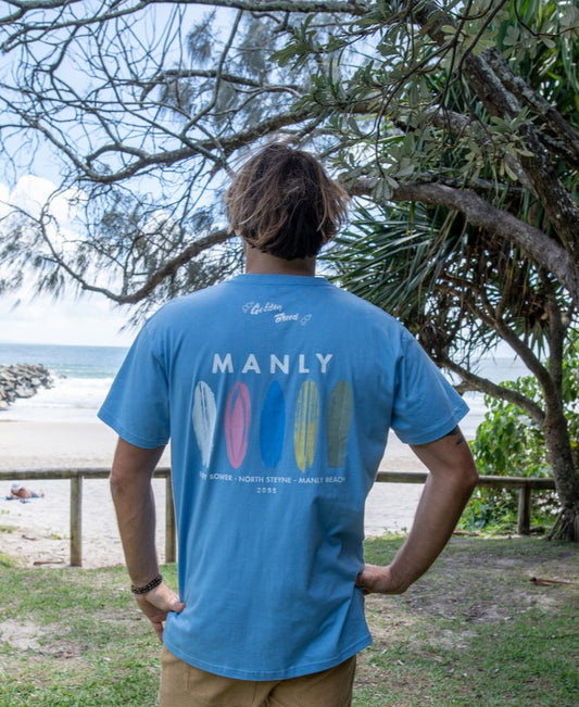 A person stands with their back to the camera, wearing a Golden Breed Manly Boards Tee in Azure Blue, gazing at a surf town beach with trees and waves in the background.