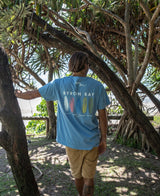 A man wearing a Golden Breed Byron Boards Tee in Azure Blue and tan shorts stands outdoors under large, leafy trees with his back to the camera.