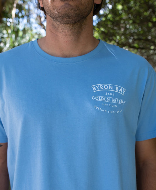 A person in the Golden Breed Byron Boards Tee in Azure Blue, featuring Surfing Since 1969 printed in white on the left chest—a classic style from Byron Bay’s iconic surf brand.