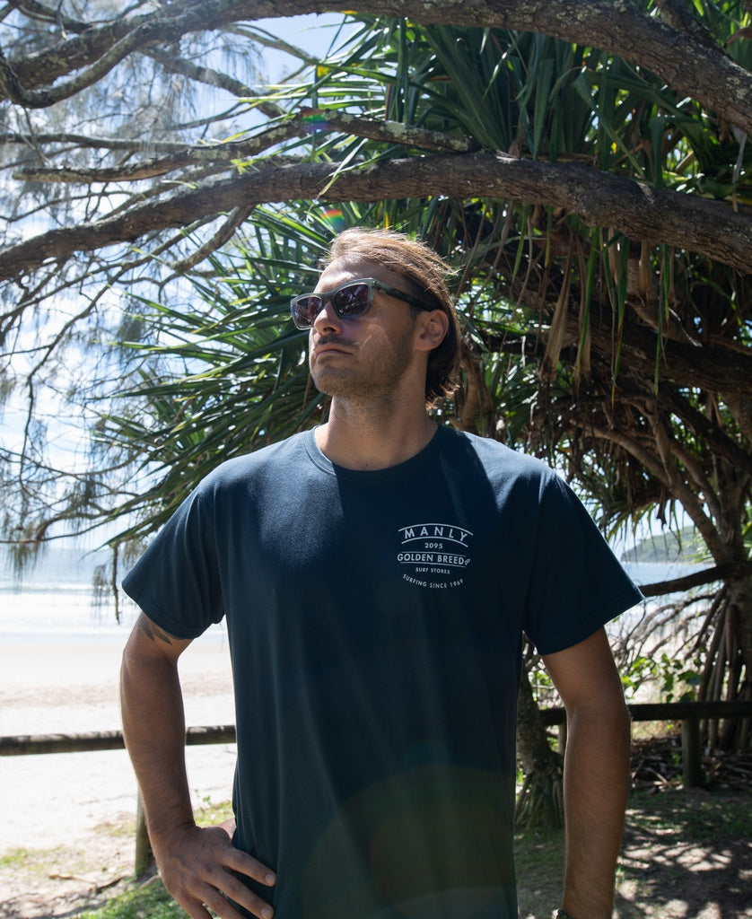 A man wearing sunglasses and a Golden Breed Manly Boards Tee in navy stands outdoors under a tree, with the beach and ocean of Surf Town visible in the background.