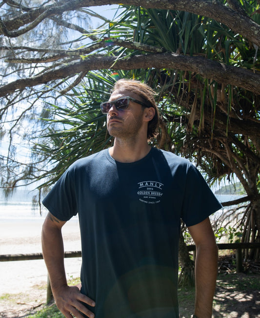A man wearing sunglasses and a Golden Breed Manly Boards Tee in navy stands outdoors under a tree, with the beach and ocean of Surf Town visible in the background.