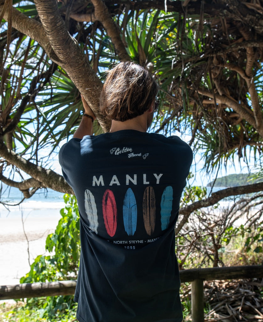 Under a leafy beach tree, a long-haired person wears the Golden Breed Manly Boards Tee in navy, crafted from soft cotton and featuring surfboard graphics with “MANLY” on the back.