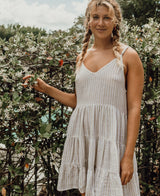 A woman with blonde braided hair wears the Golden Breed Nina Baby-doll Dress in Off White Stripe, standing before a flowering bush outdoors on a partly cloudy day.