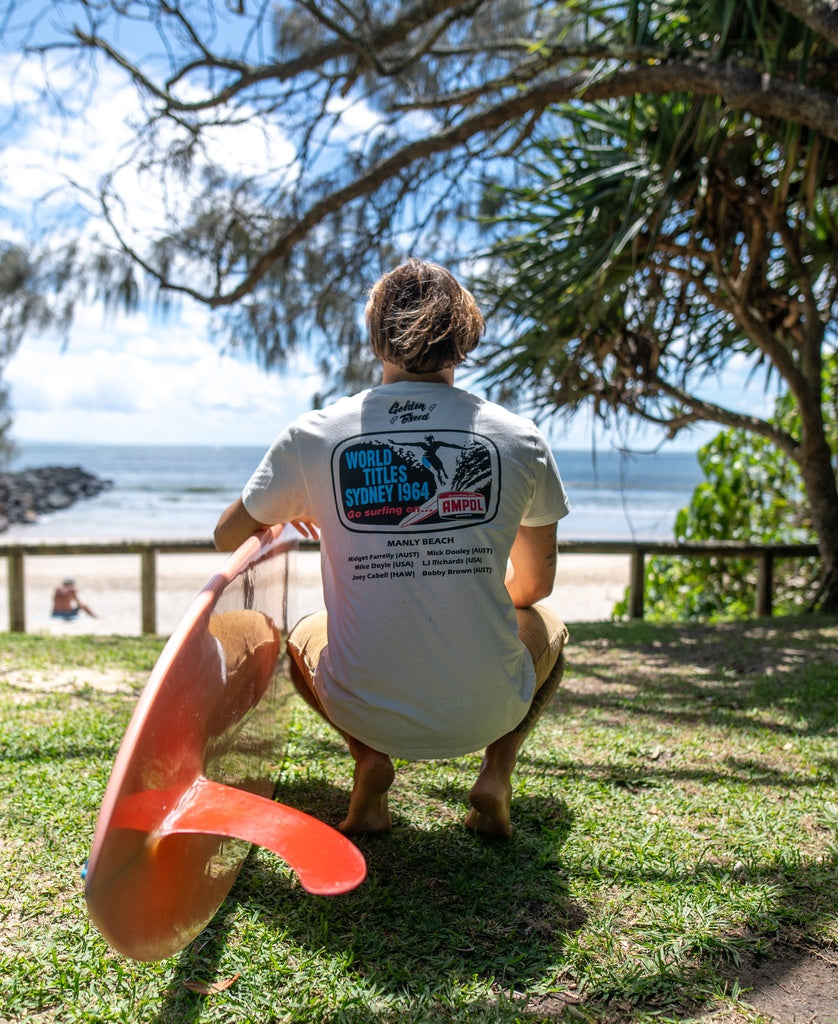 A person holding a surfboard sits on grass facing Manly Beach, wearing the Golden Breed Manly Ampol Tee | Off White, featuring an event graphic on the back.
