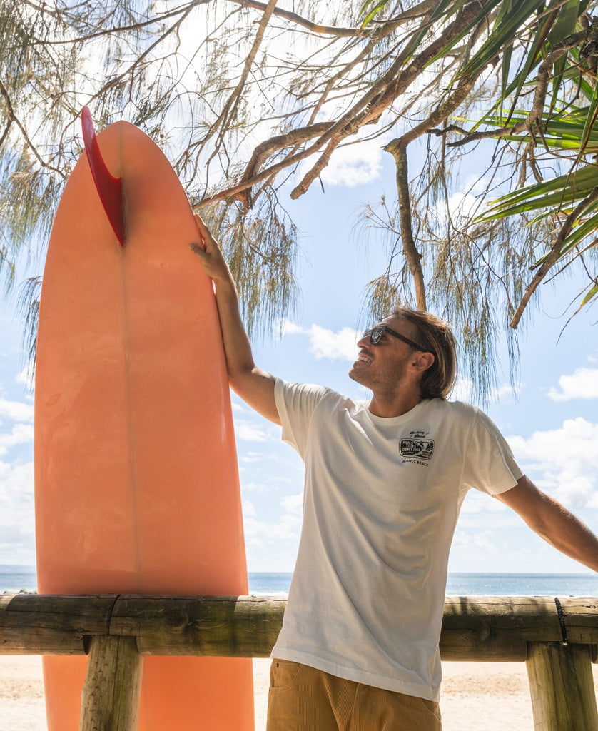 A man wearing the Golden Breed Manly Ampol Tee in Off White and sunglasses stands by a pink surfboard, smiling and looking up at Manly Beach with trees and the ocean in the background.