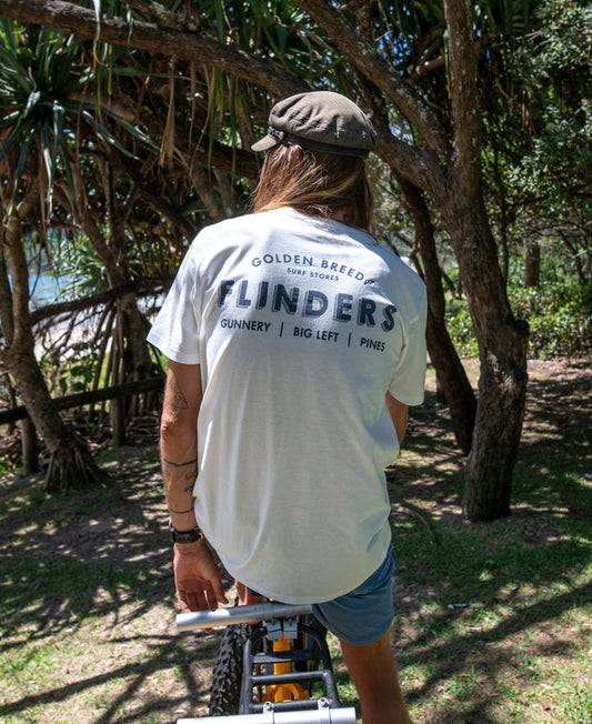 Wearing a Golden Breed Flinders Old School Tee | Off White, a person sits on a bike in a park, blending classic 100% cotton comfort with laid-back outdoor vibes.