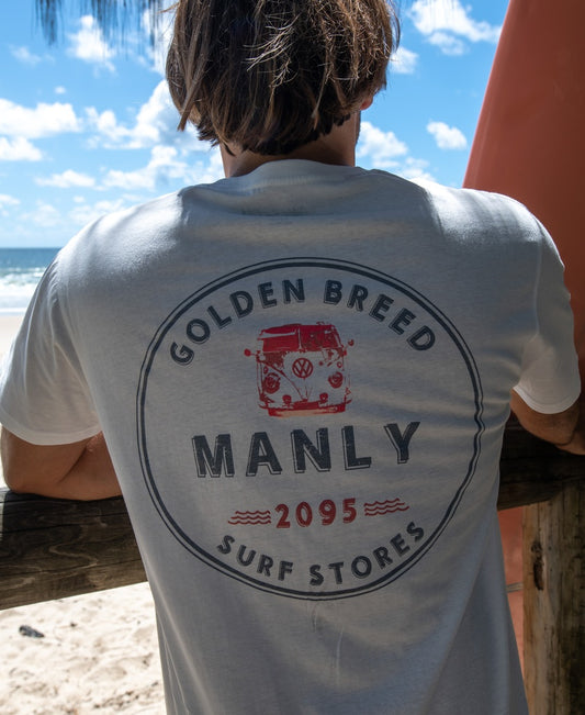 Wearing a Golden Breed Manly Kombi Tee in off white, a person stands by a wooden fence at the beach, facing the ocean.