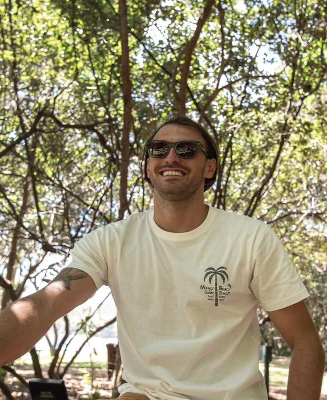 A man wearing sunglasses and a Golden Breed Manly Palm Loco Tee in Off White, featuring a palm tree design, smiles while sitting outdoors with trees and sunlight in the background.