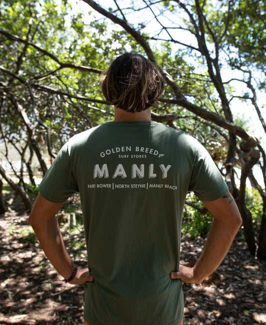 A person stands outdoors with their back to the camera, wearing a khaki Manly Old School Tee by Golden Breed—made from 100% cotton—surrounded by trees and dappled sunlight.