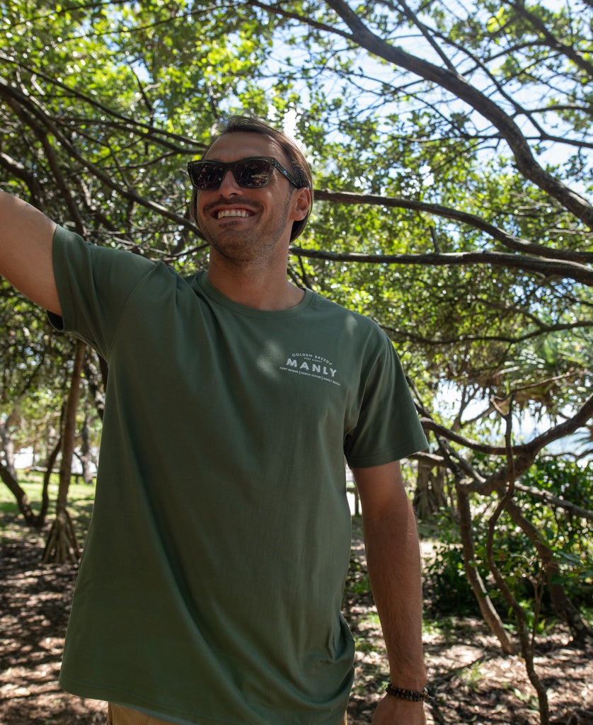 A person wearing sunglasses and a Golden Breed Manly Old School Tee in Khaki stands outdoors under leafy trees, smiling and facing slightly left.