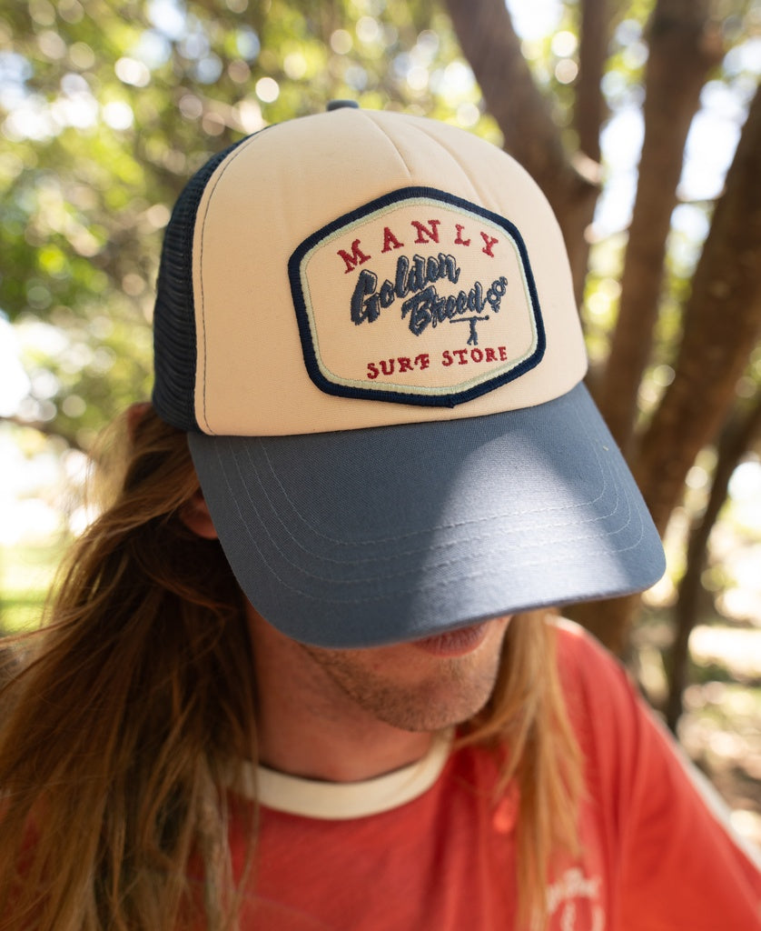Person wearing a Manly Trucker Cap | Dark Denim by Golden Breed, featuring an adjustable strap and logo on the front, outdoors with trees in the background.