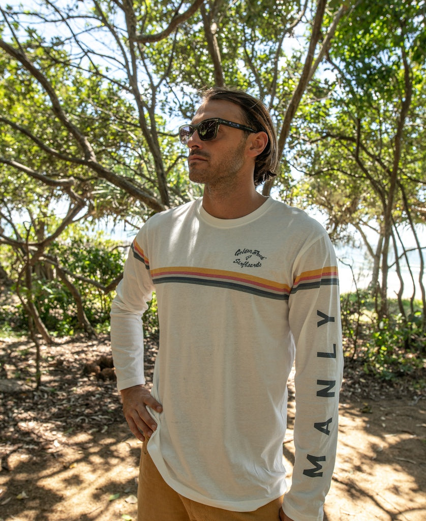 A man in sunglasses wears the Golden Breed Manly Cali Long Sleeve Tee in off white, featuring retro stripes and MANLY on the sleeve, as he stands outdoors among trees.