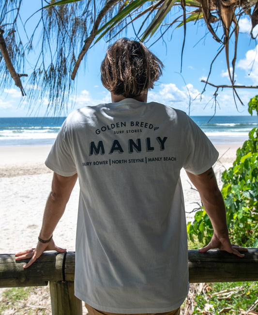 Wearing the Golden Breed Manly Old School Tee in Off White, designed in Australia from 100% cotton, a person stands at a wooden railing facing the ocean, with trees and beach behind them.