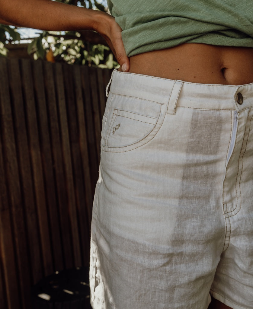 A person in a green top and Golden Breeds Lyla Short | Off White Linen stands outdoors with one hand on their hip, beside a wooden fence.