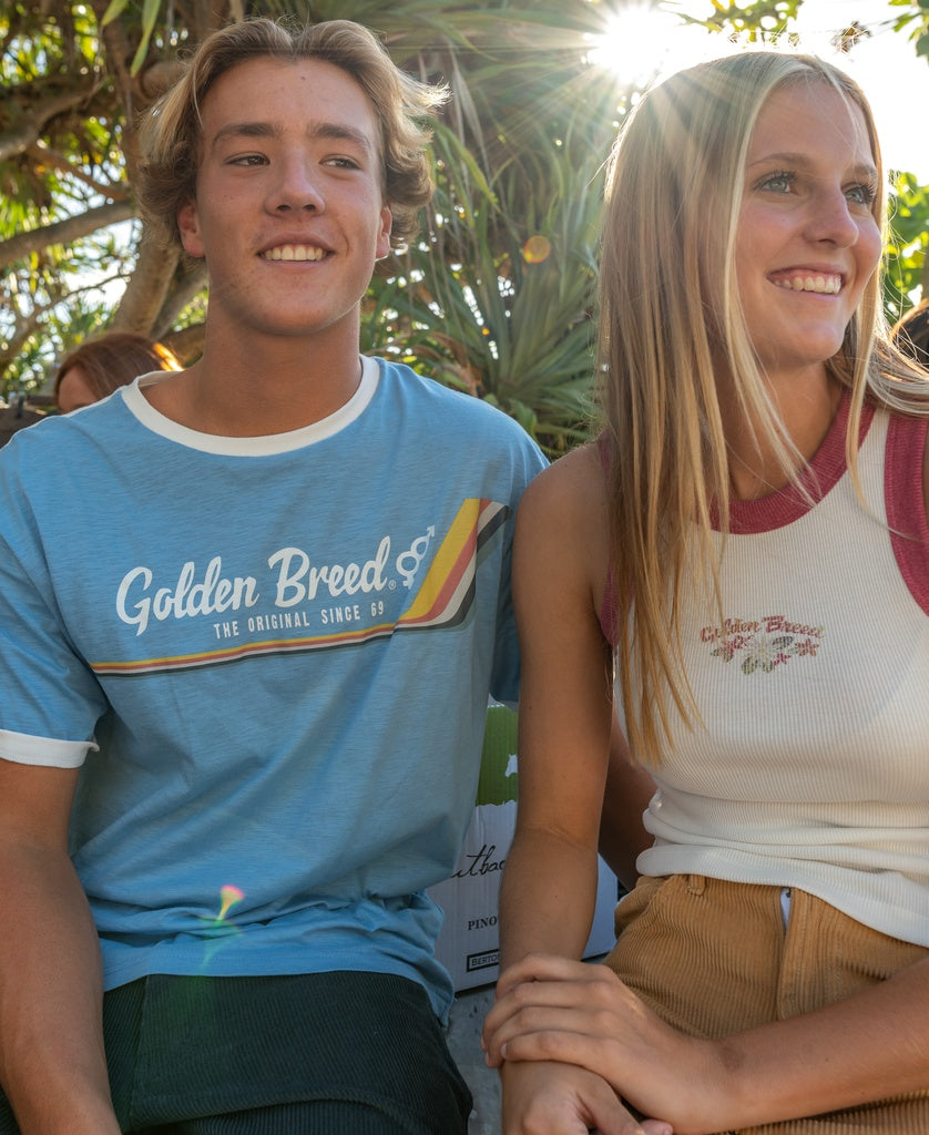 A young man and woman sit close outdoors, smiling in the sunlight. Both wear casual Golden Breed Retro Ringer Tees in Azure Blue, radiating true ’70s vibes.