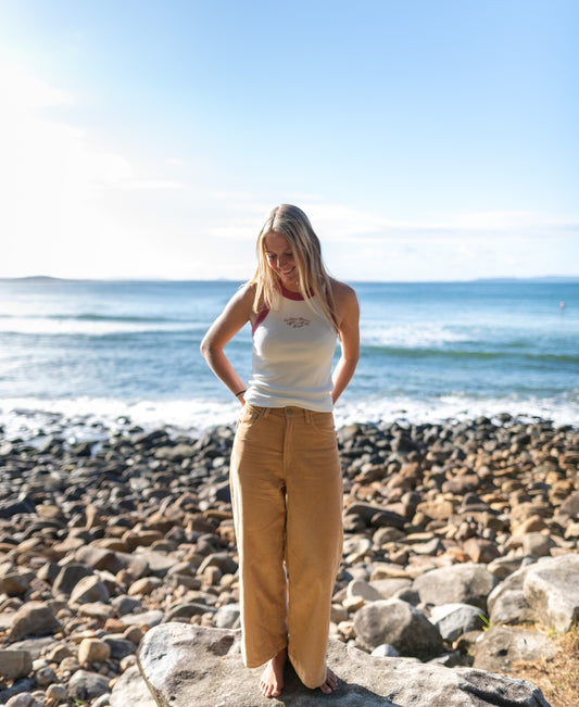 A woman stands barefoot on a rocky beach, gazing down, wearing Golden Breed’s Jenny Cord Pants in Camel with the ocean and blue sky behind her.