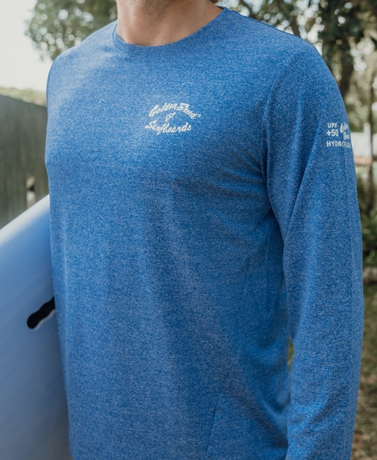 A person wearing the Golden Breed Pebbles L/S Rash Shirt in Blue Marle with UPF 50+ protection stands outdoors holding a surfboard; brand text appears on the chest and sleeve.