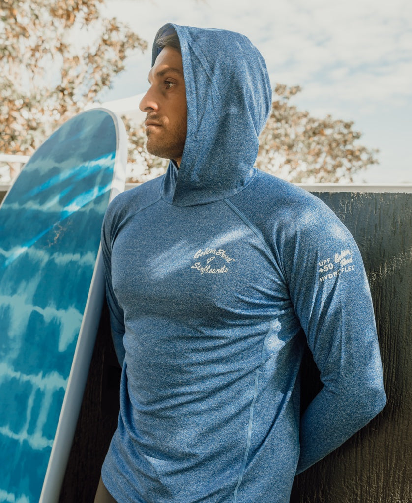 A man wearing the Golden Breed Rapper Hoodie Rash Shirt in Blue Marle stands in front of a blue surfboard and wooden fence, with trees in the background.