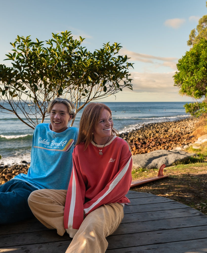 Two people relax on a wooden deck by the ocean in slouchy retro style, both wearing Golden Breed’s Retro Rag Top | Sky Marle. Smiling, they gaze right as waves, rocks, trees, and an embroidered GB logo complete the breezy scene.