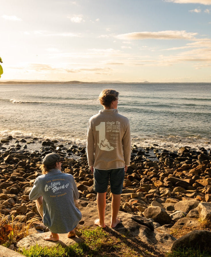 Two people in casual wear, including the Golden Breed Fin 1/4 Zip Rag Top in Mocha Marle, stand and sit on grass by a rocky shore, gazing at the ocean under a partly cloudy sky.