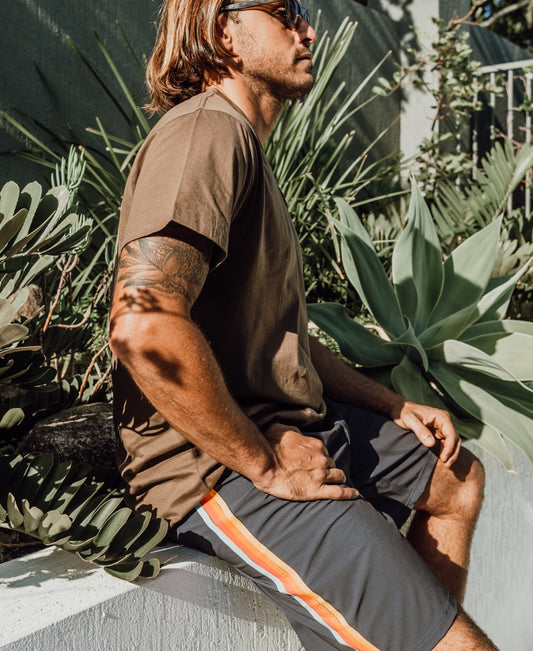 A man in sunglasses, a brown t-shirt, and Golden Breed’s Long Island Boardie | Wash Black shorts relaxes on a low wall among green plants in the sun, channeling that laid-back 60’s California surf vibe.