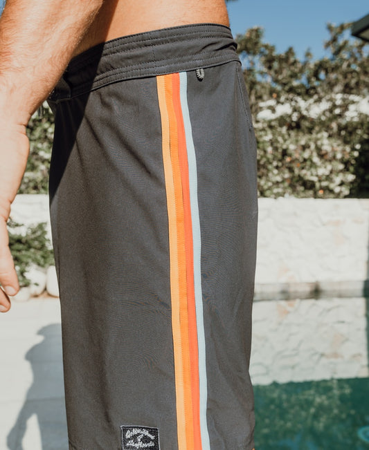 A person stands by the pool in Golden Breed’s Long Island Boardie | Wash Black shorts, which feature orange, red, and blue vertical stripes on the sides for a retro 60’s California surf vibe.