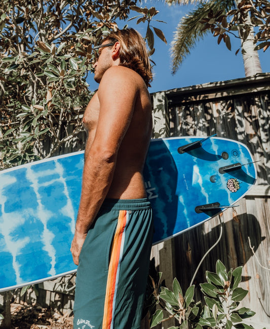 A shirtless man stands outdoors holding a blue surfboard, surrounded by greenery and a wooden fence, wearing Golden Breeds Long Island Elastic Waist Boardie in Ivy Green, designed with 4-way stretch for comfort.