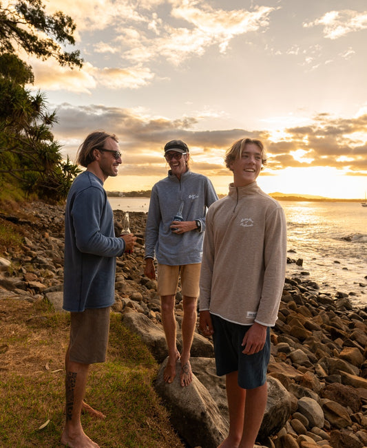 Three men in Golden Breed Fin 1/4 Zip Rag Top | Mocha Marle stand on a rocky shoreline at sunset, smiling and talking as their French Terry shirts catch the evening light, with the ocean and a boat in the background.