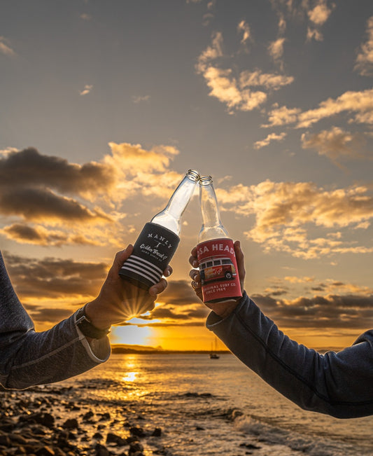 Two people clink beer bottles with Golden Breed Noosa Stubby Kombi | Wash Red holders at sunset by the water, clouds overhead and the sun low on the horizon.