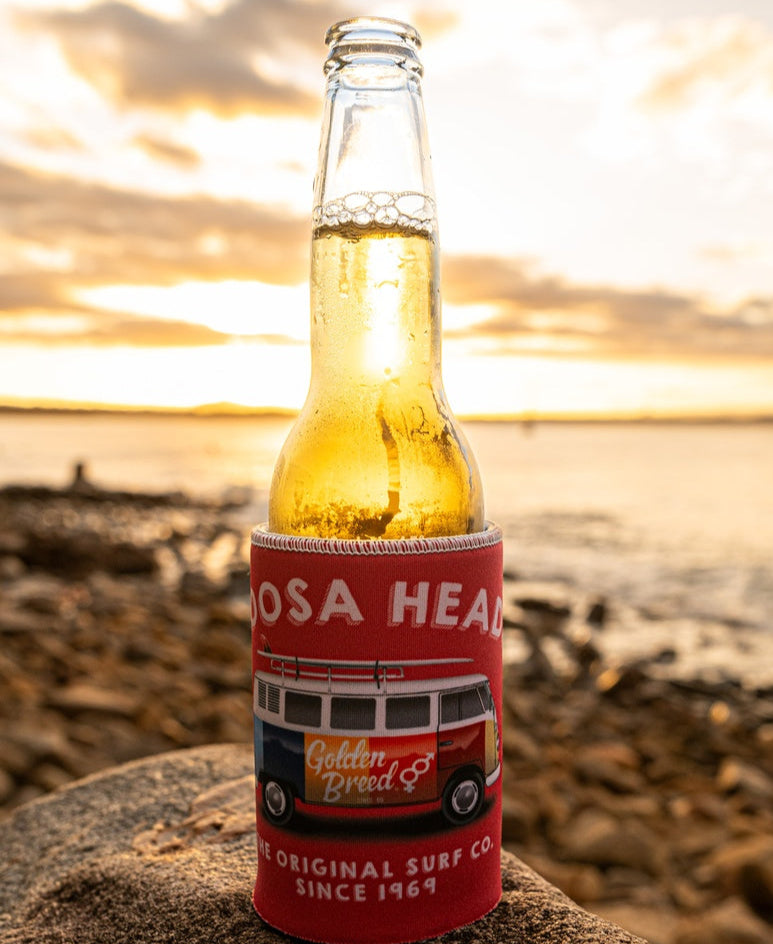 A clear glass beer bottle rests on a rock by the beach at sunset in a Golden Breed Noosa Stubby Kombi | Wash Red holder, crafted from durable neoprene with a vibrant sublimated print.