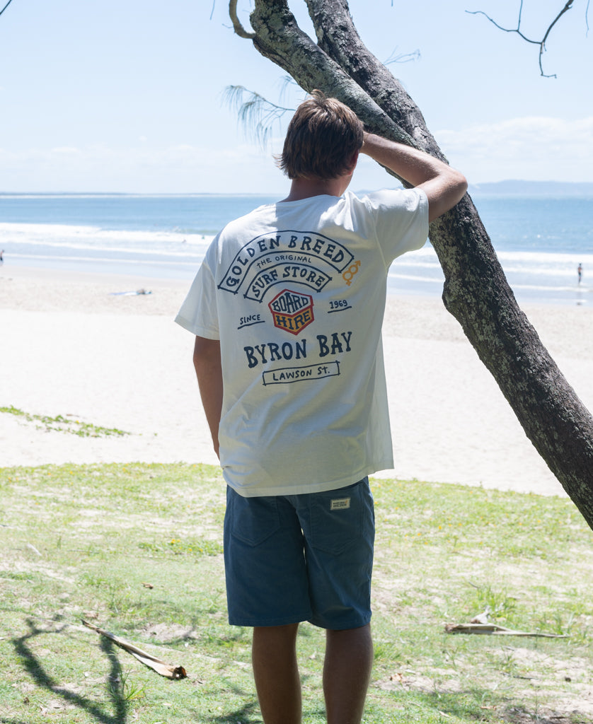 Man standing on grassy shore near ocean wearing Byron Herring Tee | Off White with surf-themed graphic on back, blue shorts, and leaning against tree under clear sky