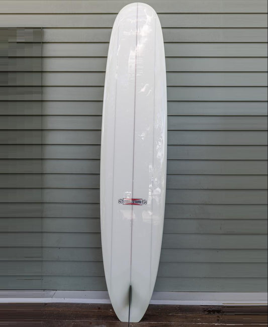 The Gordon & Smith G&S X Thomas Longboard 96, a white, single-fin board with a pointed nose for modern performance and noseriding, stands upright against a gray paneled wall.