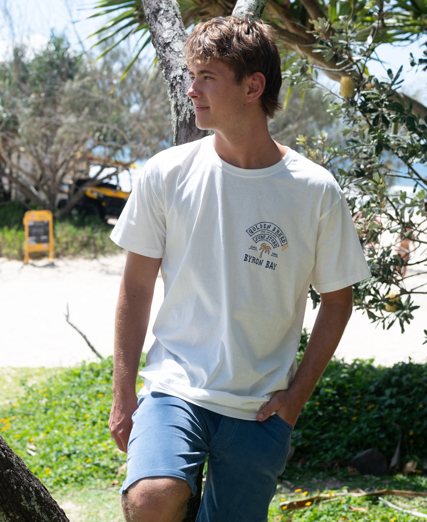 A young man wearing a Byron Herring Tee | Off White stands outdoors near trees in a sunny, relaxed beachside setting, capturing casual Australian surfwear style.