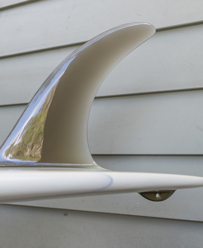 Close-up of a G&S X Thomas Longboard 94 surfboard fin by Gordon & Smith, attached to the bottom of a white longboard ideal for noseriding, set against a light gray wooden wall.
