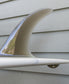Close-up of a G&S X Thomas Longboard 94 surfboard fin by Gordon & Smith, attached to the bottom of a white longboard ideal for noseriding, set against a light gray wooden wall.