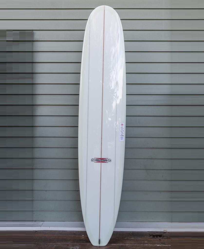 The Gordon & Smith G&S X Thomas Longboard 94 with a single wooden stringer stands upright against a light gray horizontal panel wall.