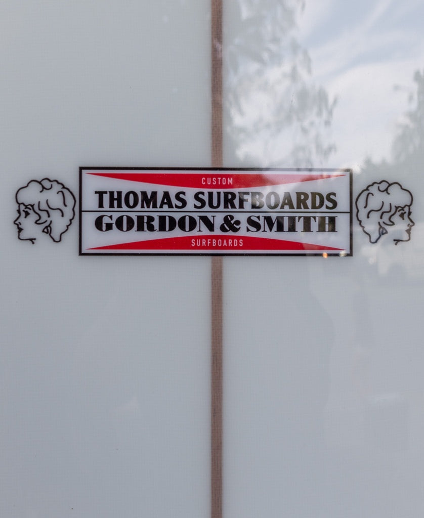 Close-up of the G&S X Thomas Longboard 94 by Gordon & Smith, featuring a logo with two illustrated profile heads inspired by classic longboard noseriding style.