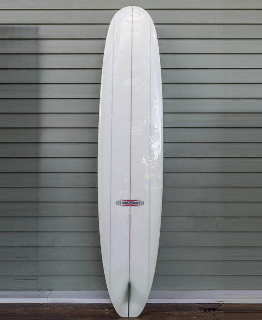 The Gordon & Smith G&S X Thomas Longboard 94 features a pointed nose and rounded tail, shown upright against a gray horizontal-paneled wall—a modern board designed for noseriding performance.