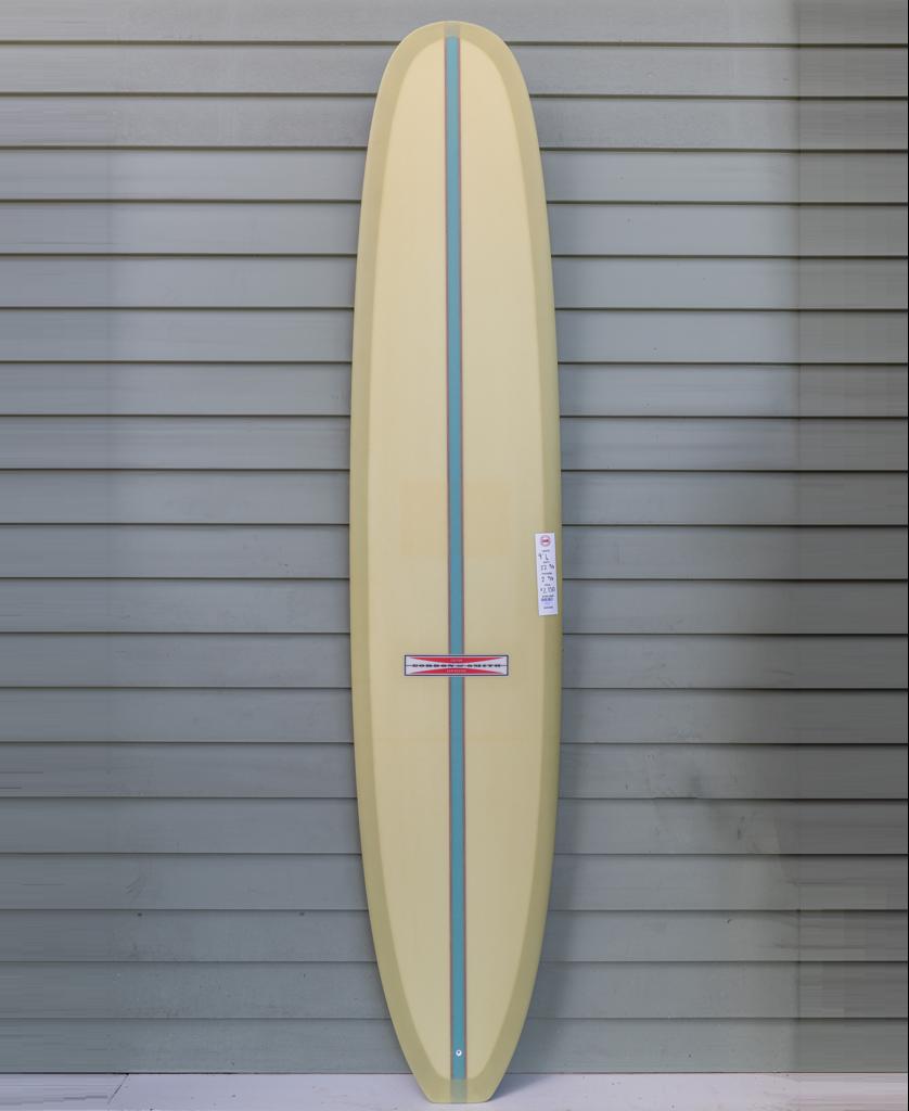 The Gordon & Smith G&S Log Sahara Sun 94 longboard surfboard stands upright against a gray paneled wall, featuring a classic yellow finish and a blue and red center stripe.