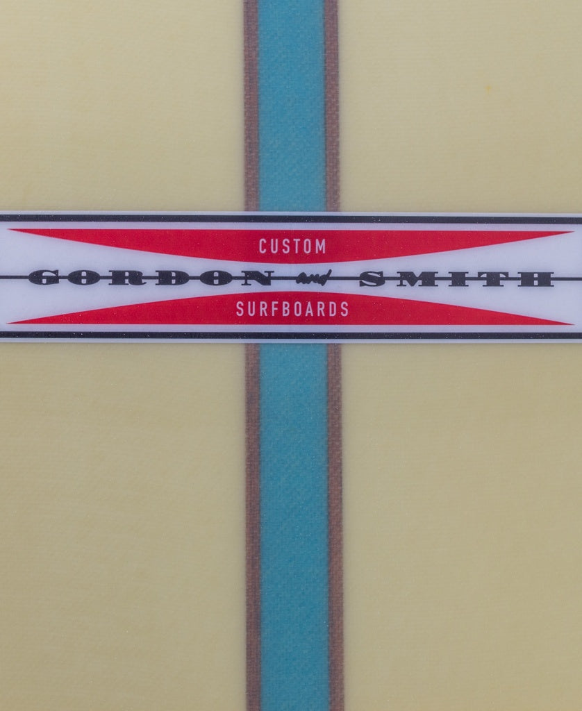 Close-up of a Gordon & Smith G&S Log Sahara Sun 94 logo with a blue vertical stripe on a light yellow traditional longboard.