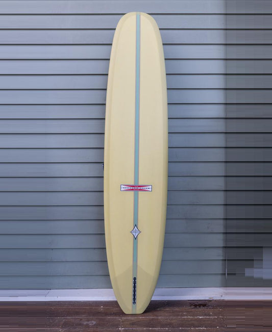 The Gordon & Smith G&S Log Sahara Sun 94 longboard, featuring a yellow finish with a blue center stripe and geometric logo, stands upright against light gray horizontal siding.