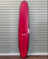 A Gordon & Smith G&S Log Cardinal 98 longboard surfboard stands upright against a light gray horizontal siding wall.