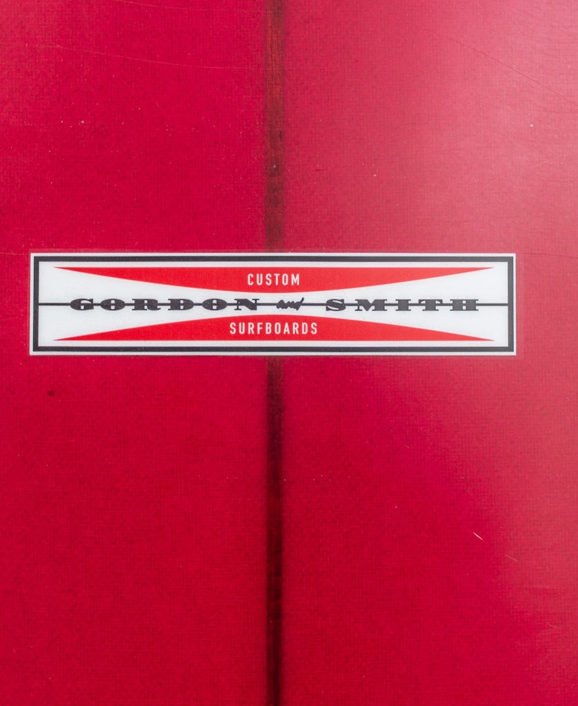 A close-up of the G&S Log Cardinal 98 red longboard surfboard by Gordon & Smith, featuring the brand’s logo label at the center.