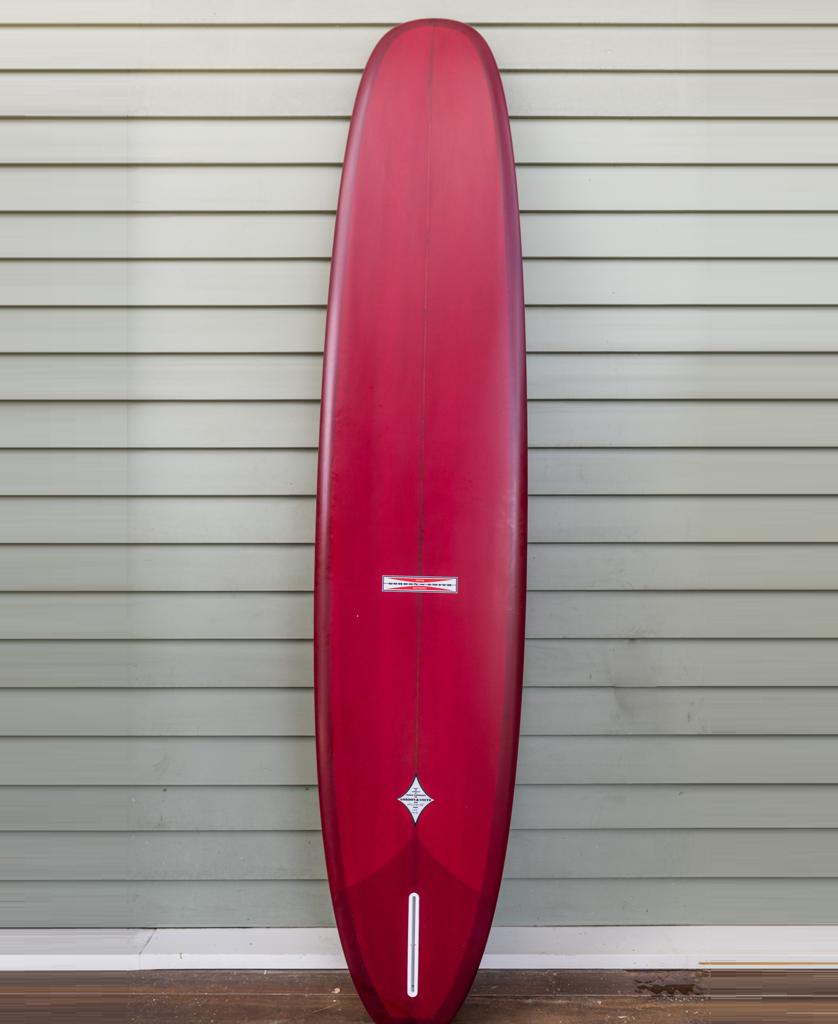 The Gordon & Smith G&S Log Cardinal 98 longboard in red stands upright against a light gray, horizontally paneled wall.