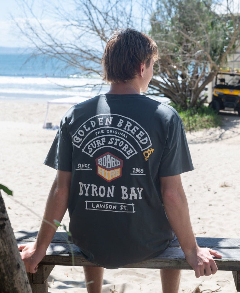 Man sitting on a bench facing the beach wearing a Byron Herring Tee | Wash Black with a retro Golden Breed surf store graphic on the back, bright sunny day at the shore.