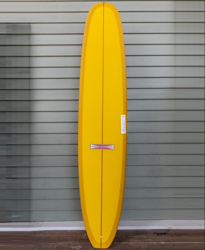 The Gordon & Smith G&S Log Gold Fusion 94, a yellow traditional longboard with classic rails, stands upright against a light gray paneled wall.
