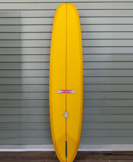 The Gordon & Smith G&S Log Gold Fusion 94 traditional longboard surfboard, featuring classic rails and a yellow finish, stands upright against a light gray, horizontally-paneled wall.